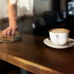 Xícara de café com espuma sobre um pires branco em uma mesa de madeira, enquanto uma pessoa limpa o balcão ao fundo em um ambiente aconchegante de cafeteria em Goiânia