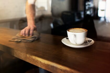 Xícara de café com espuma sobre um pires branco em uma mesa de madeira, enquanto uma pessoa limpa o balcão ao fundo em um ambiente aconchegante de cafeteria em Goiânia